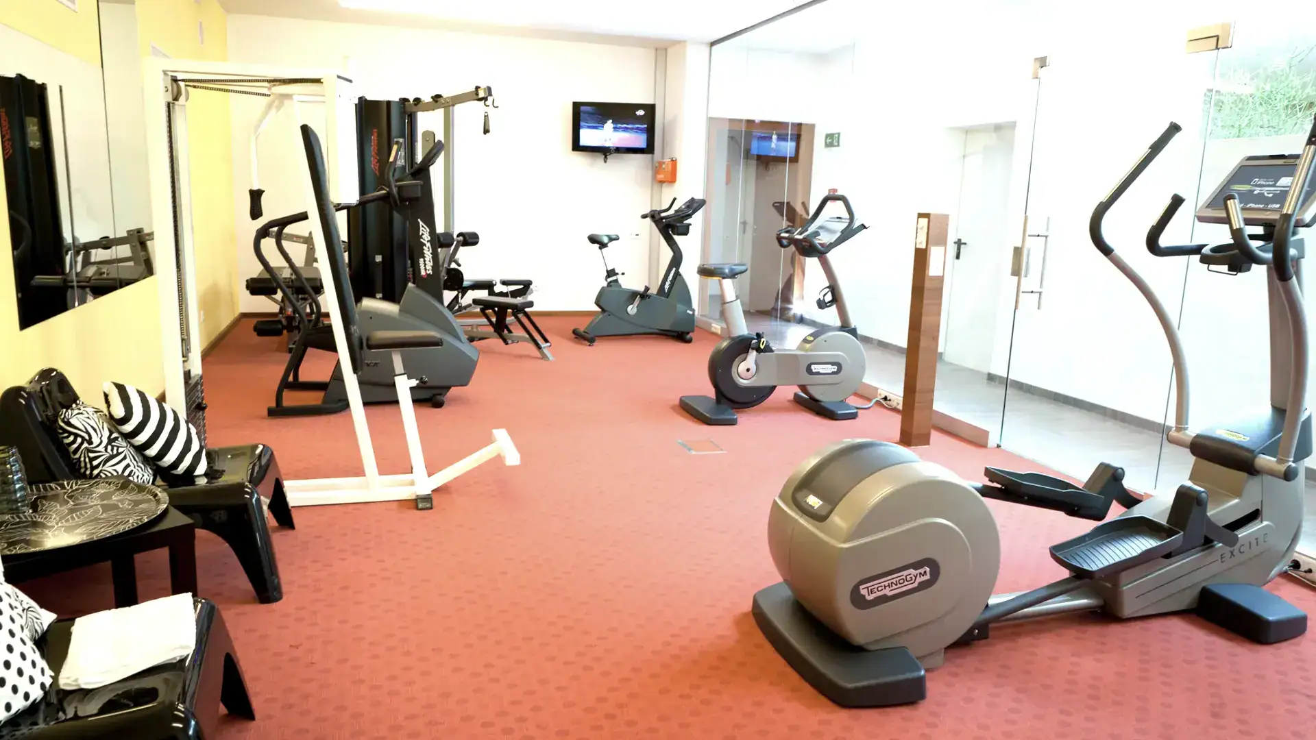 Fitnessstudio mit verschiedenen Geräten und roten Teppichboden.