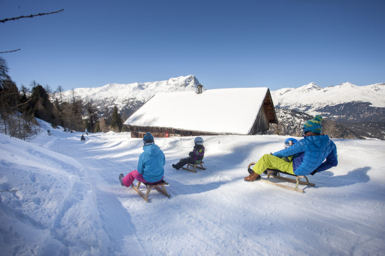 Familie rodelt auf schneebedecktem Hang mit Berghütte.