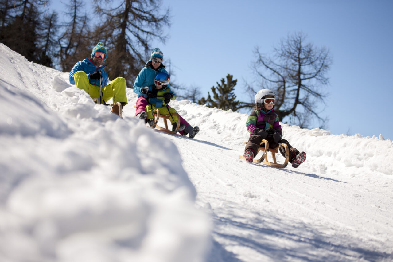 Familie beim Rodeln im Schnee unter blauem Himmel.